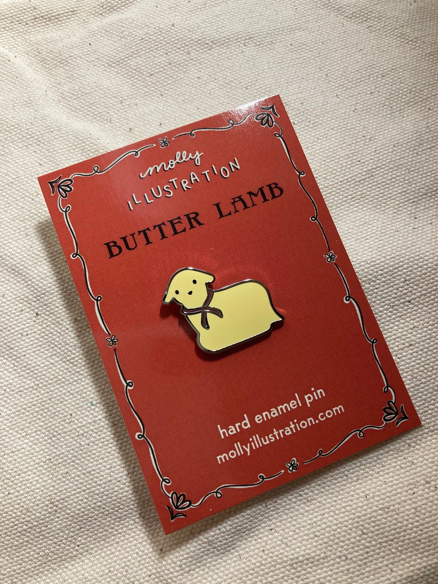 a hard enamel pin of a yellow butter lamb with red ribbon around it's neck. It is on a red Molly Illustration backer card.