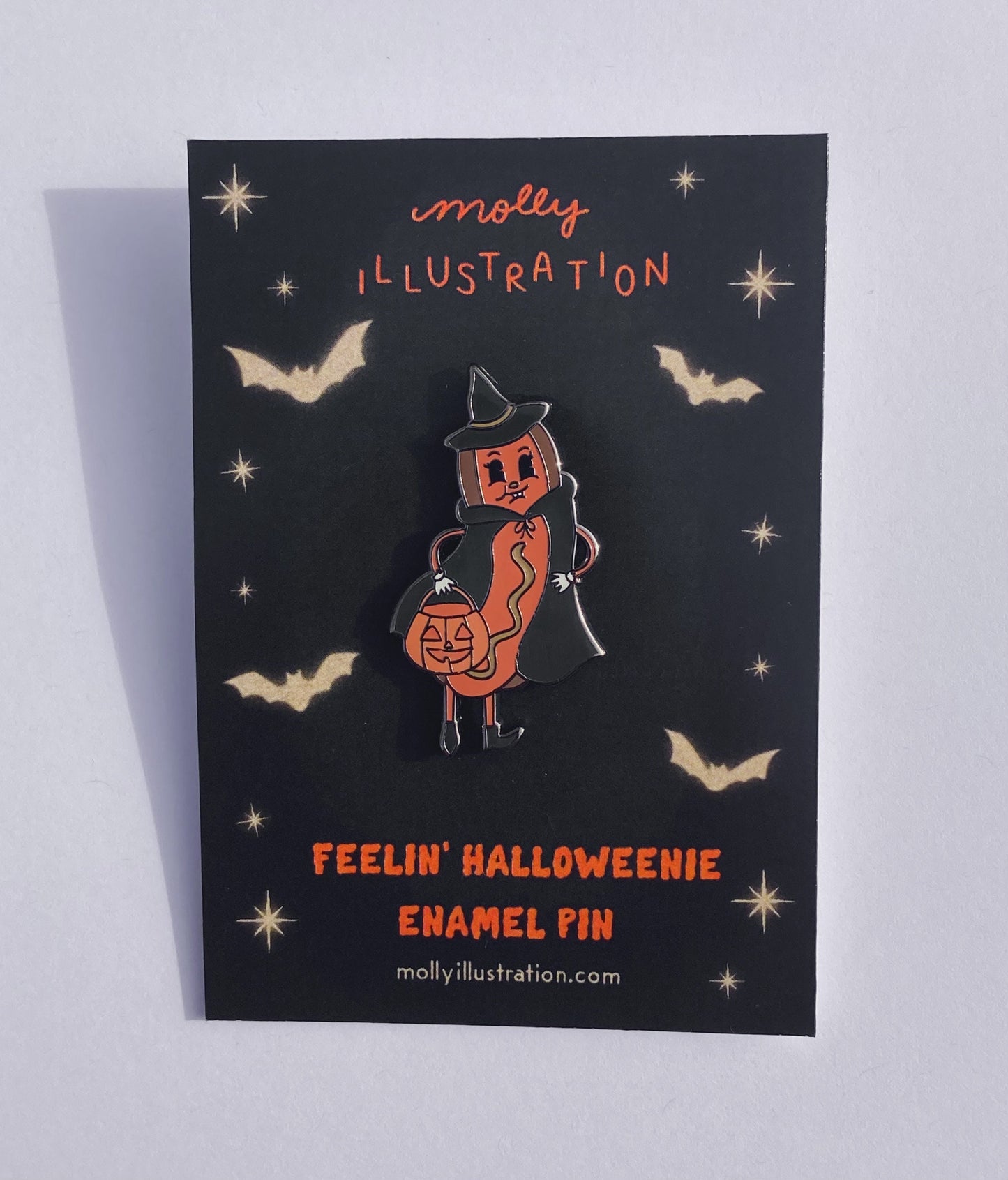 A hard enamel pin of a Halloween Hot Dog dressed as a witch and holding a jack o lantern bucket. It is on a molly illustration backer card.