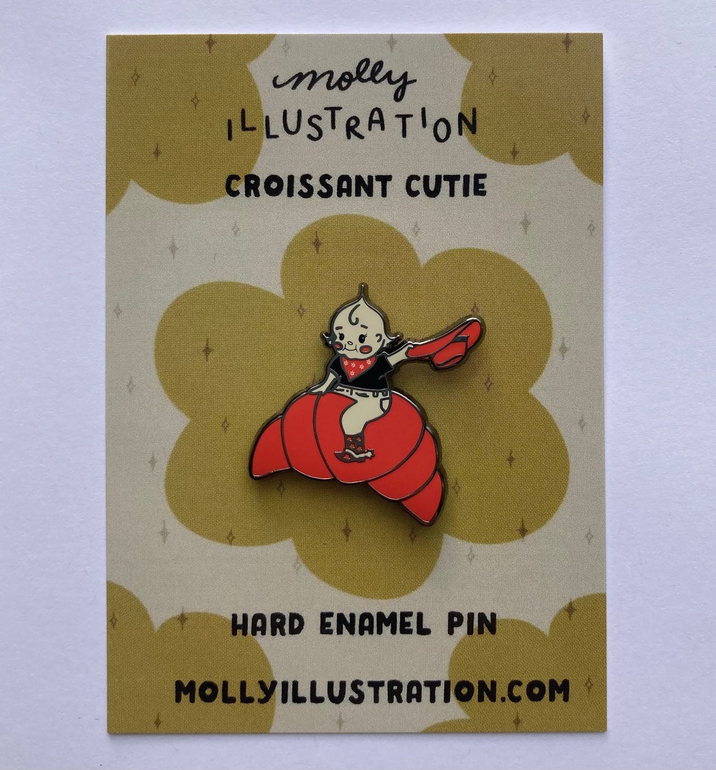 A hard enamel pin of a cartoon croissant cutie cowgirl. the background is a Molly Illustration backer card