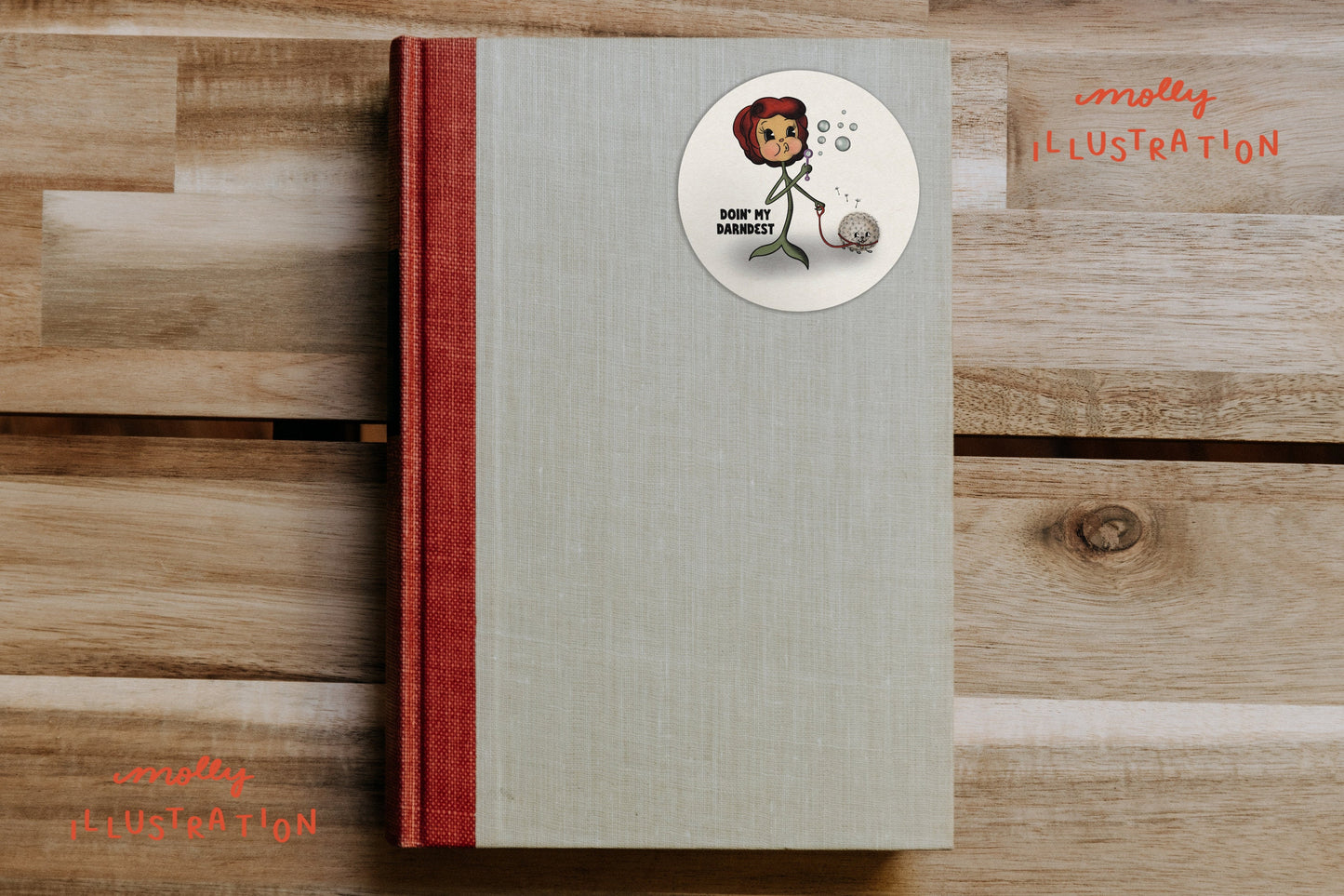 A round vinyl sticker of a cartoon flower blowing bubbles, walking her dandelion that reads "Doin my Darndest" stuck onto a blank book cover.