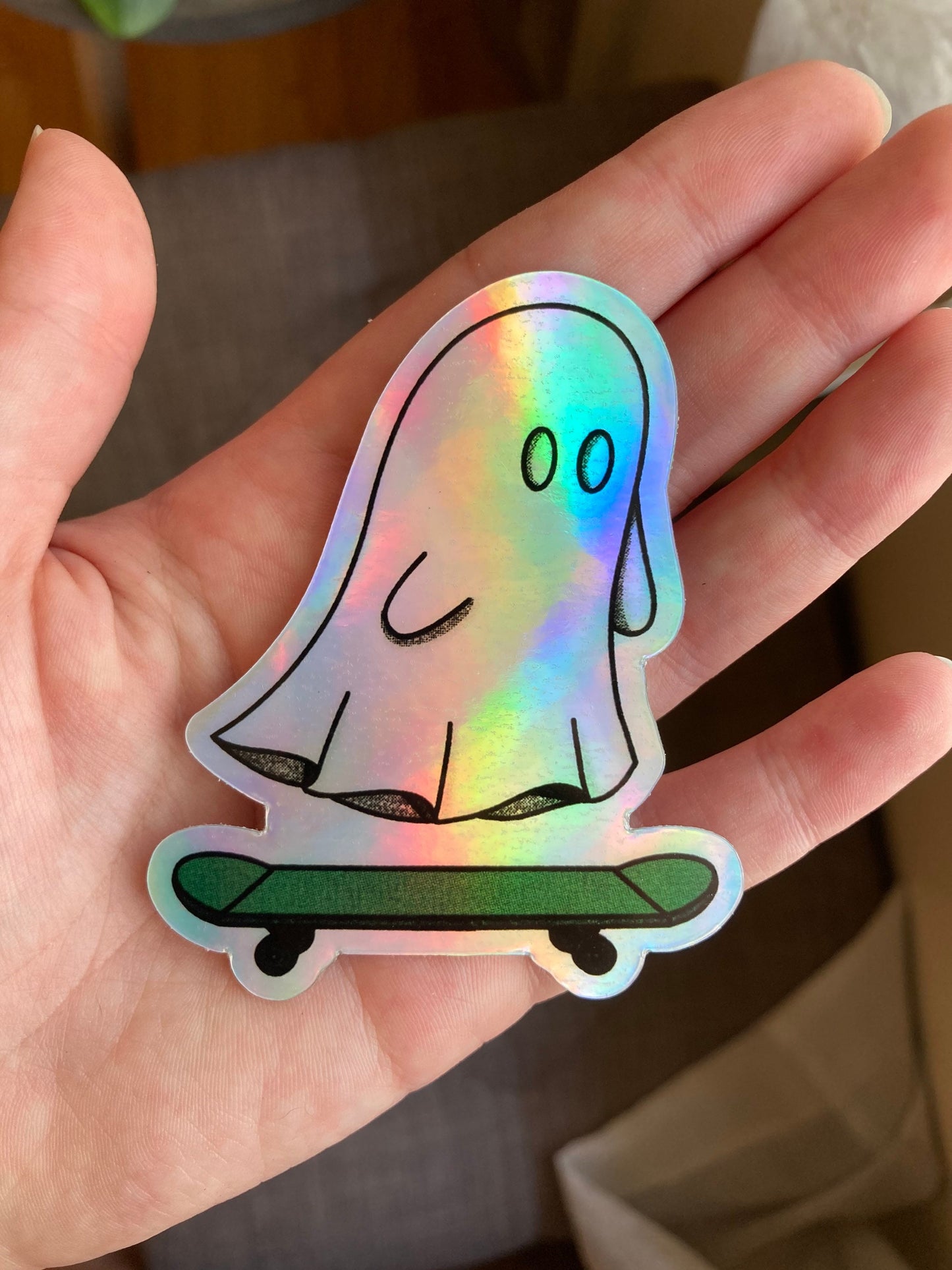 a hand holding an illustrated, waterproof, holographic vinyl sticker of a ghost riding a skateboard