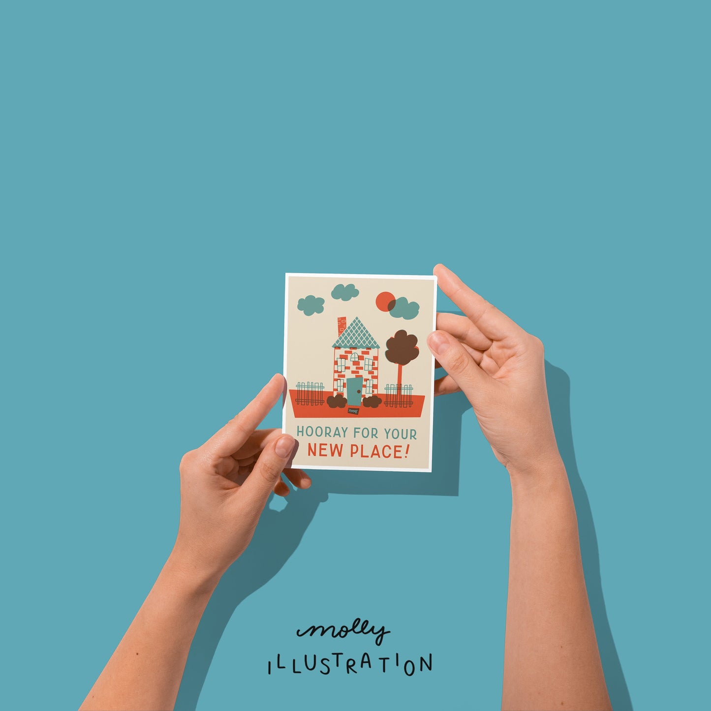 hands holding an illustrated vertical greeting card that shows a red and blue house scene. it reads "hooray for your new place!"