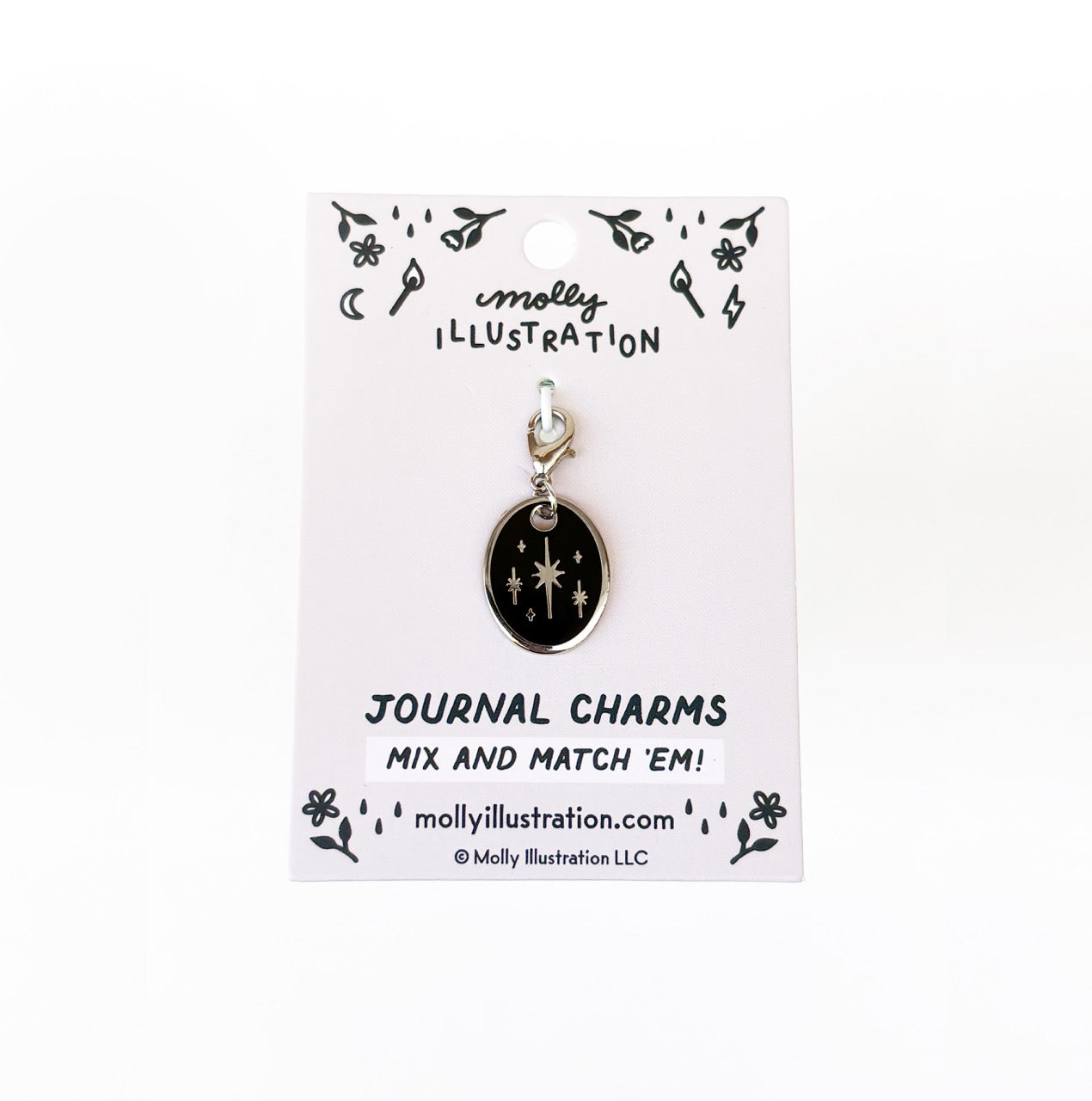 A silver and black journal charm with stars on it. Behind it is a branded backer card for the product
