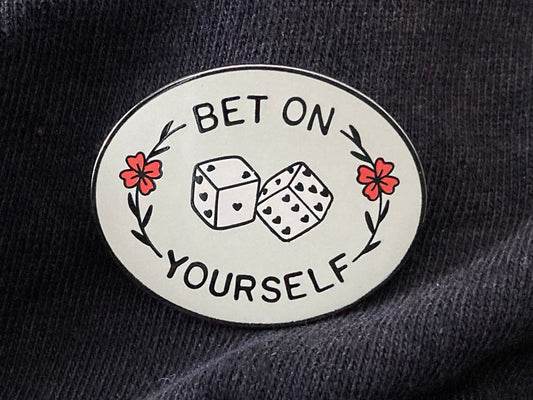 A hard enamel pin that reads "Bet on Yourself." with a set of dice in the center. It is over a gray fabric background.