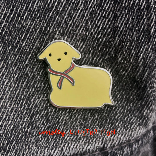 a hard enamel pin of a yellow butter lamb with red ribbon around it's neck. it is over a gray denim background.