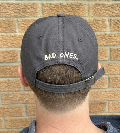 Person wearing a gray cap with 'BAD ONES.' text against a brick wall.