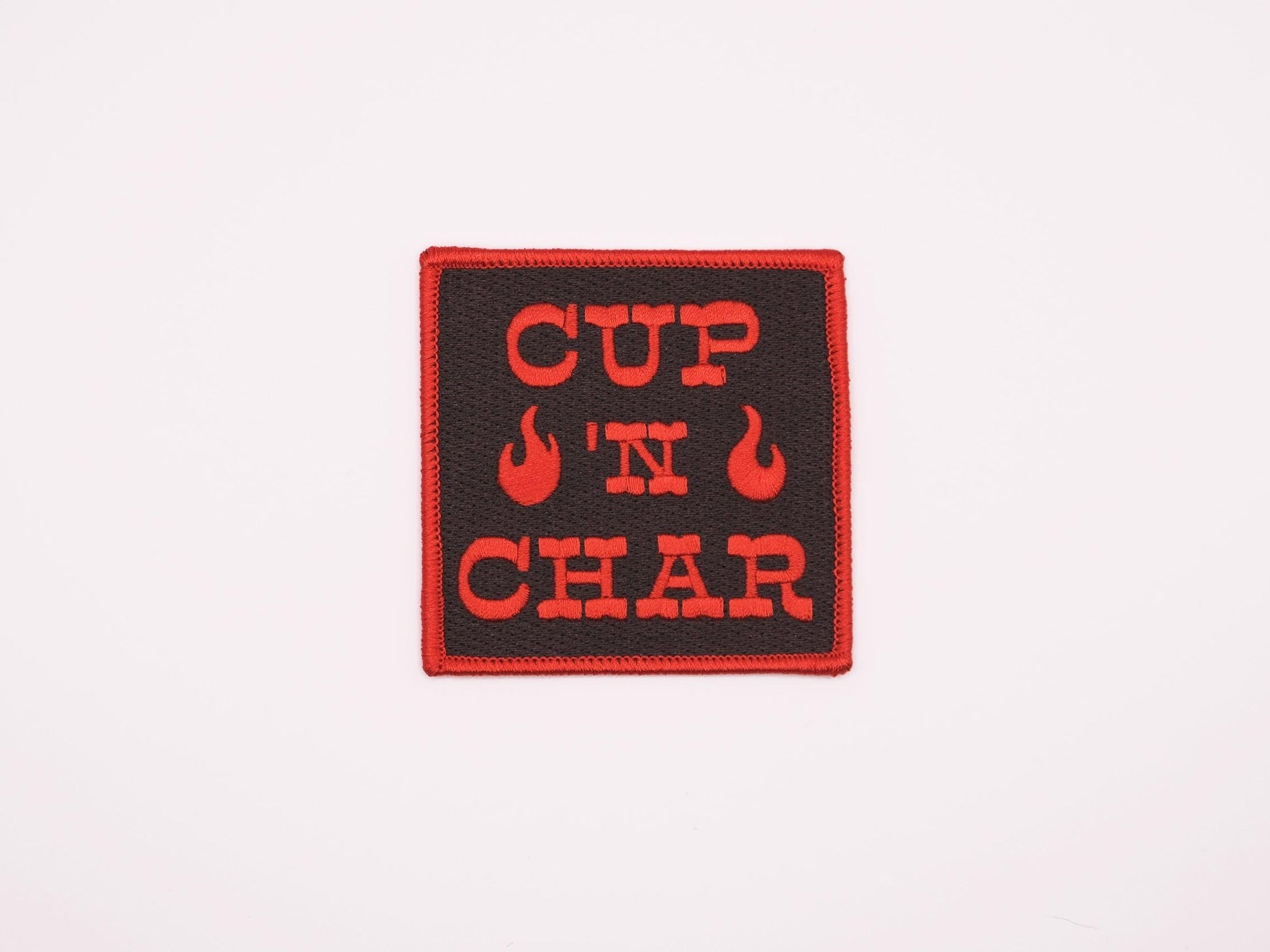 A black square embroidered iron on patch that reads "CUP 'N CHAR" in red letters. there are two red flames beside the "'N" and there is a red merrowed border.