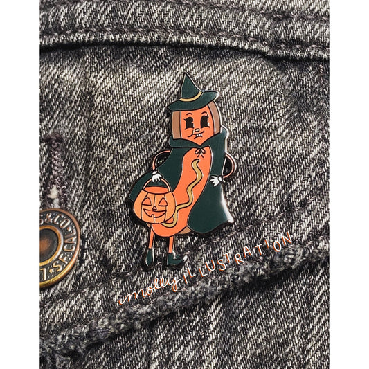 A hard enamel pin of a Halloween Hot Dog dressed as a witch and holding a jack o lantern bucket. It is over a gray denim background.