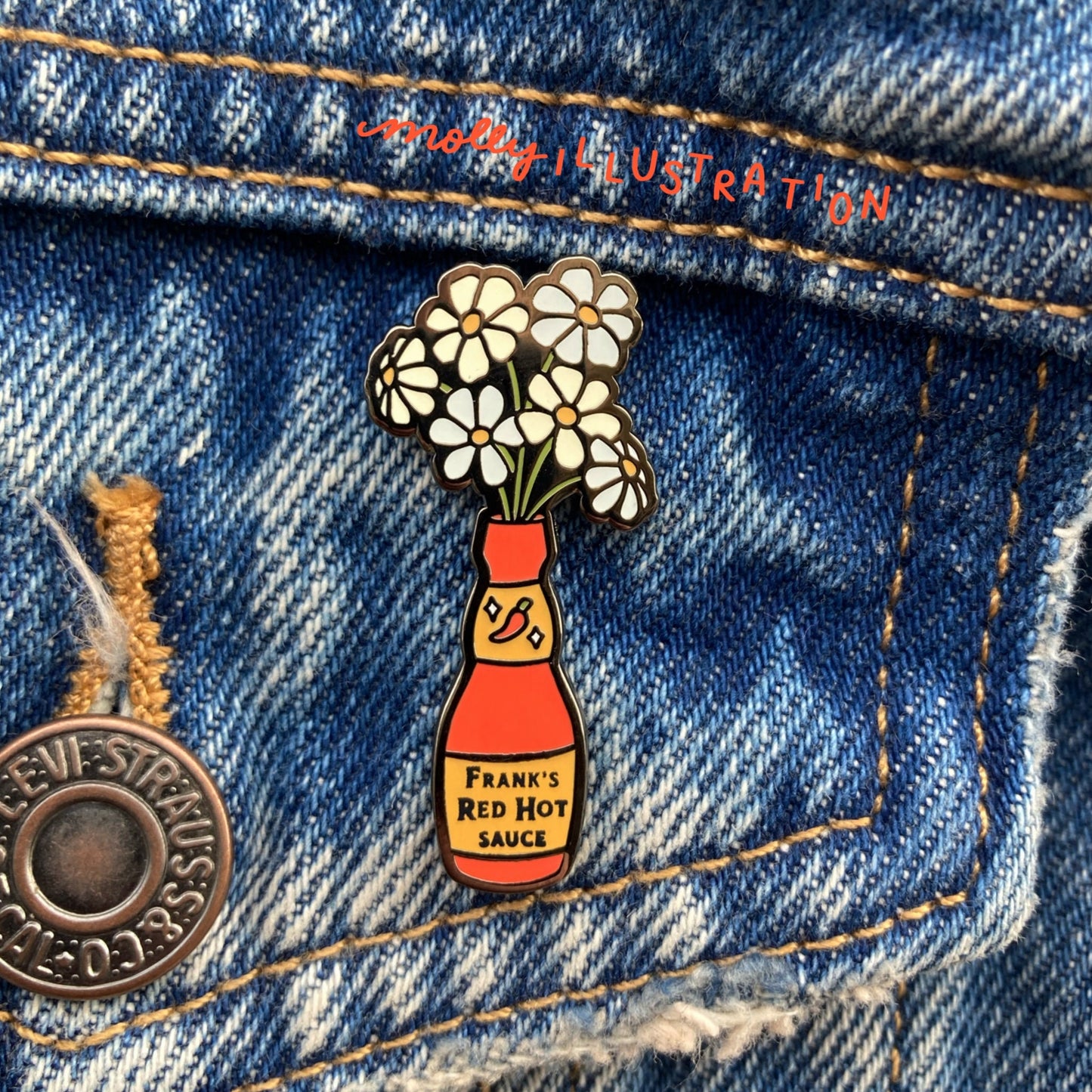 A hard enamel pin of a hot sauce bottle that is being used as a vase for daisies. It is over a denim background