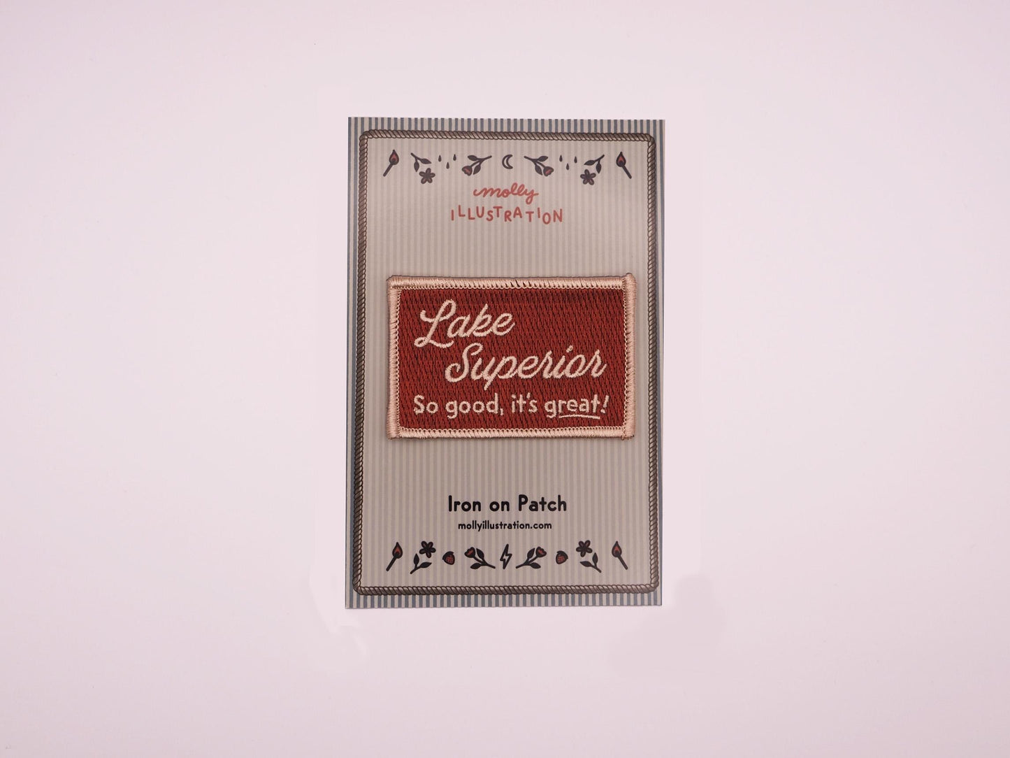 a burgundy rectangular embroidered iron on patch that reads "Lake Superior, So good, it's great!" in off white lettering packaged on a striped Molly Illustration backer card