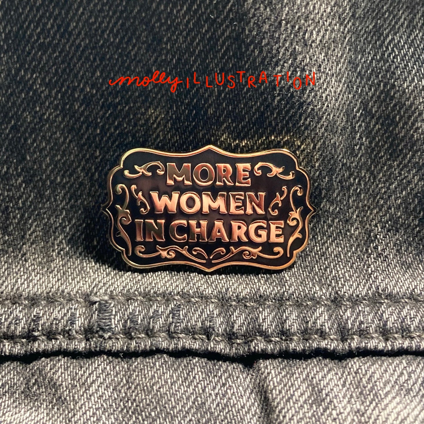 A hard enamel pin that reads "More Women in Charge." it is black and gold. It is over a gray denim background