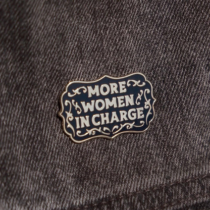 A hard enamel pin that reads "More Women in Charge." it is black and gold. It is over a gray denim background.