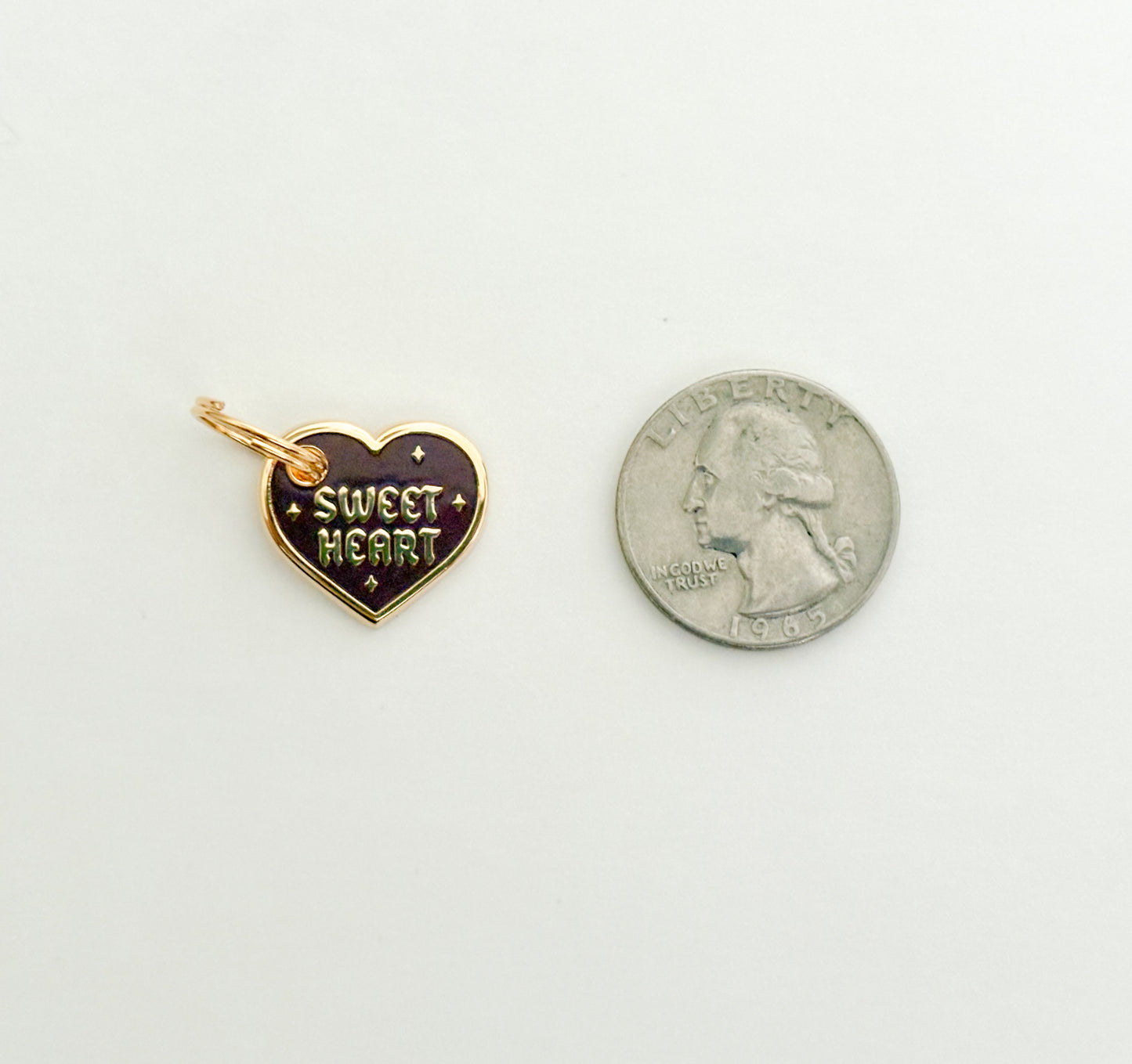 A burgundy and gold colored collar charm that reads "Sweet Heart" next to a quarter on a white background