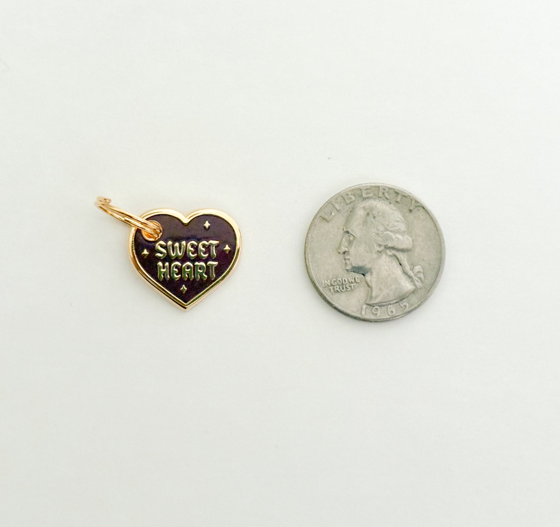 A burgundy and gold colored collar charm that reads "Sweet Heart" next to a quarter on a white background