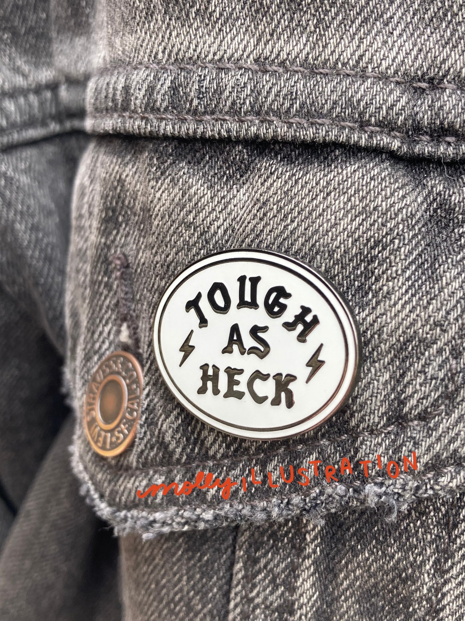 A hard enamel pin that reads "Tough as Heck." with a set of lightning bolts on the side. It is over a background of grey denim.