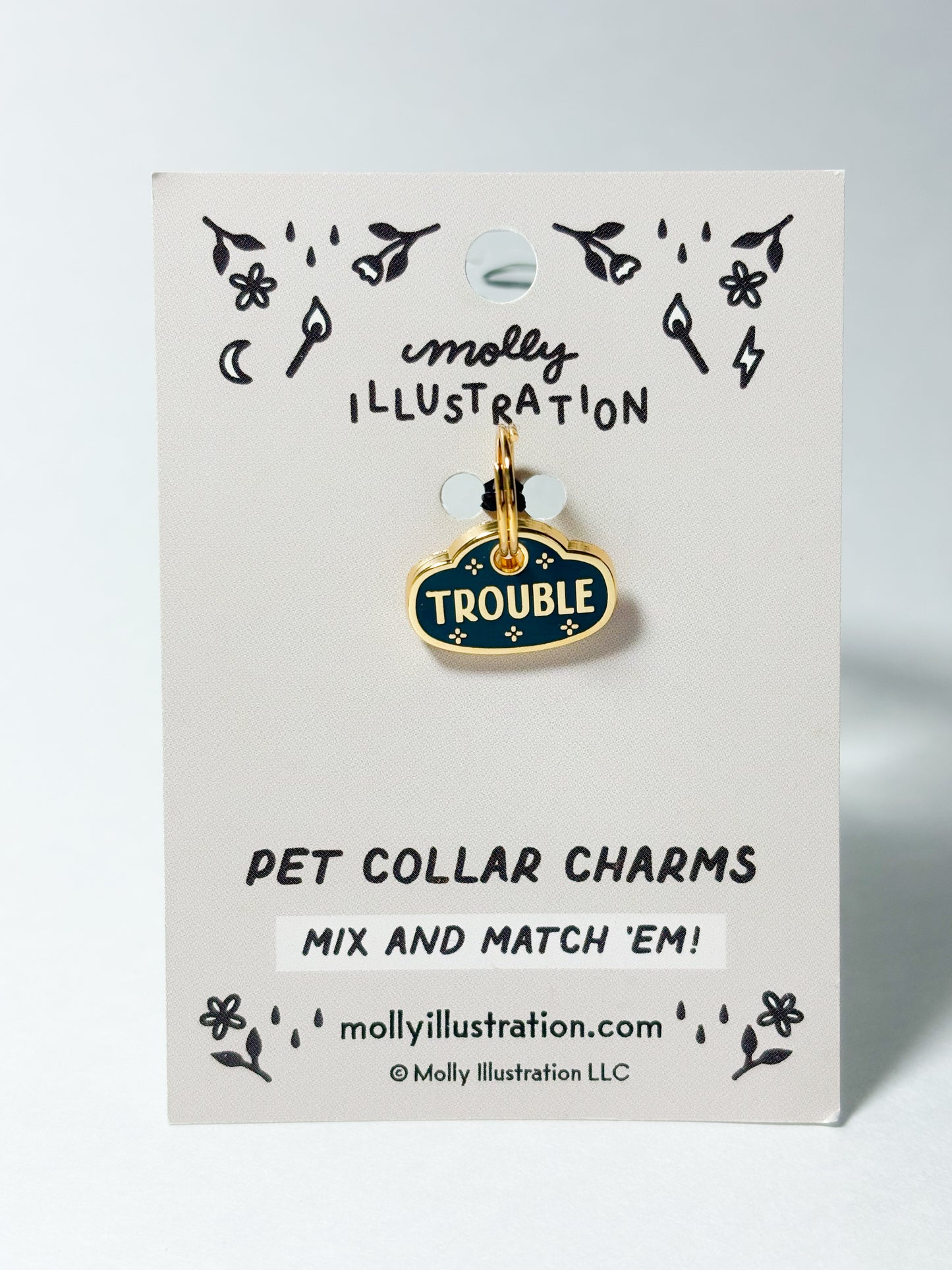 A dark teal and gold colored pet collar charm that reads 'TROUBLE' on a Molly Illustration branded card.