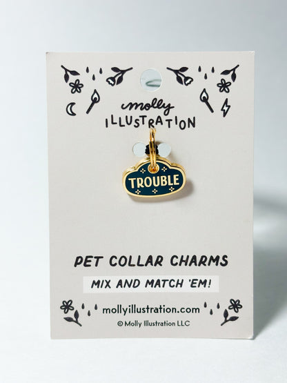 A dark teal and gold colored pet collar charm that reads 'TROUBLE' on a Molly Illustration branded card.