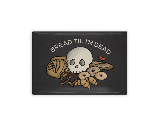 a rectangular refrigerator magnet showing the front and back. the front is dark grey and has a cartoon of a skull on a pile of various breads and reads "Bread Til I'm Dead" above the drawing