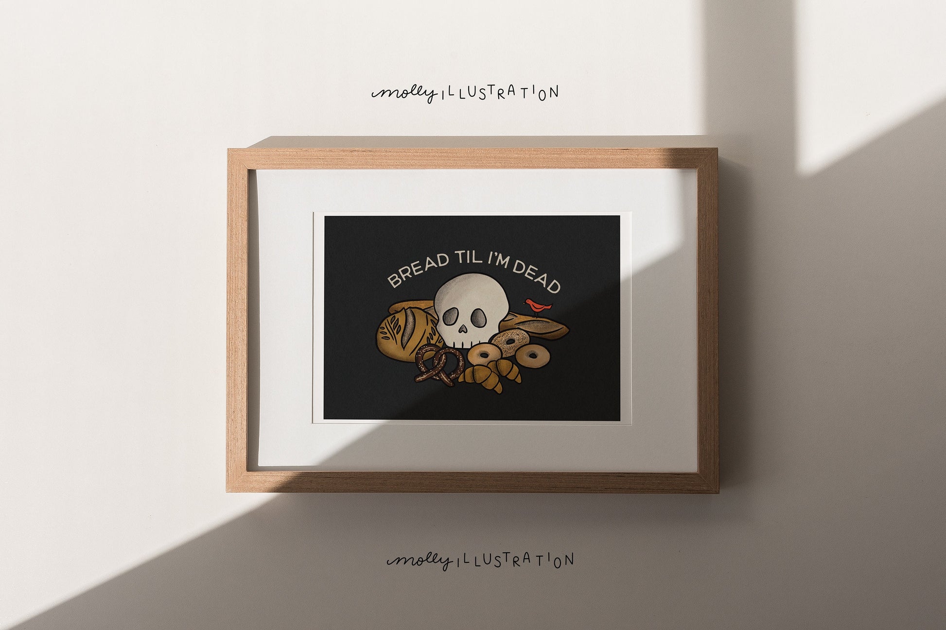 An illustrated art print of various cartoon breads and pastries with a skull sitting on top of them. A small red bird sits nearby and is eating a loaf of bread. Above it reads “Bread til I’m Dead.”