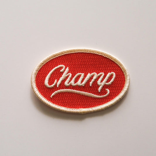 A red embroidered iron on patch that reads “Champ" in off white script lettering.