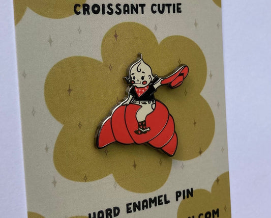 A hard enamel pin of a cartoon croissant cutie cowgirl. the background is a Molly Illustration backer card