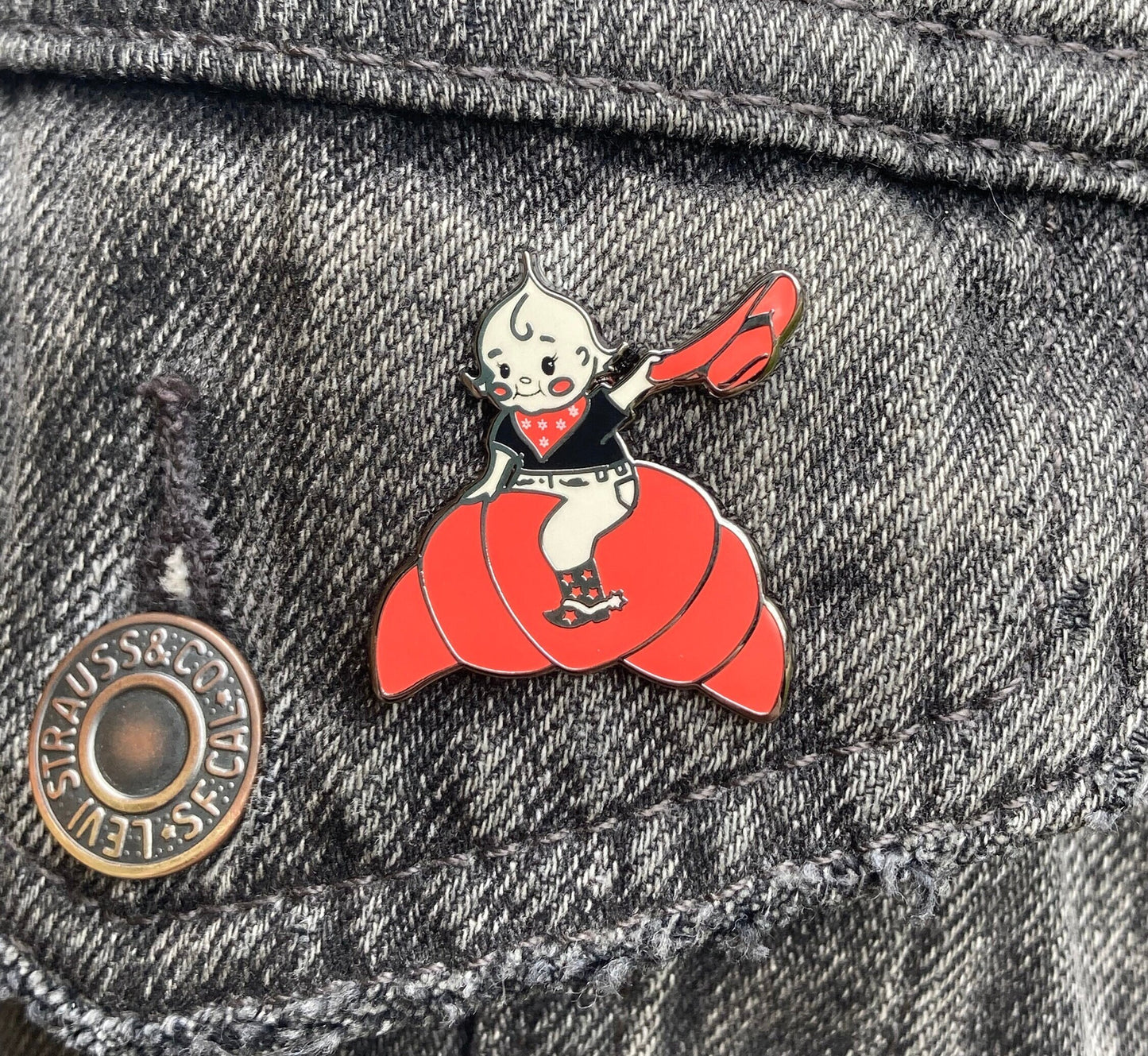 A hard enamel pin of a cartoon croissant cutie cowgirl. It is over a gray denim background