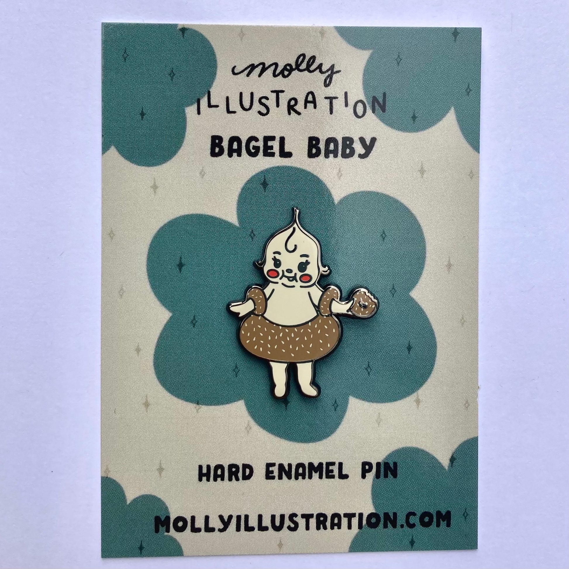 A hard enamel pin of a cartoon bagel baby. It is packaged on a Molly Illustration backer card.