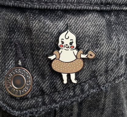 A hard enamel pin of a cartoon bagel baby. It is over a gray denim background.