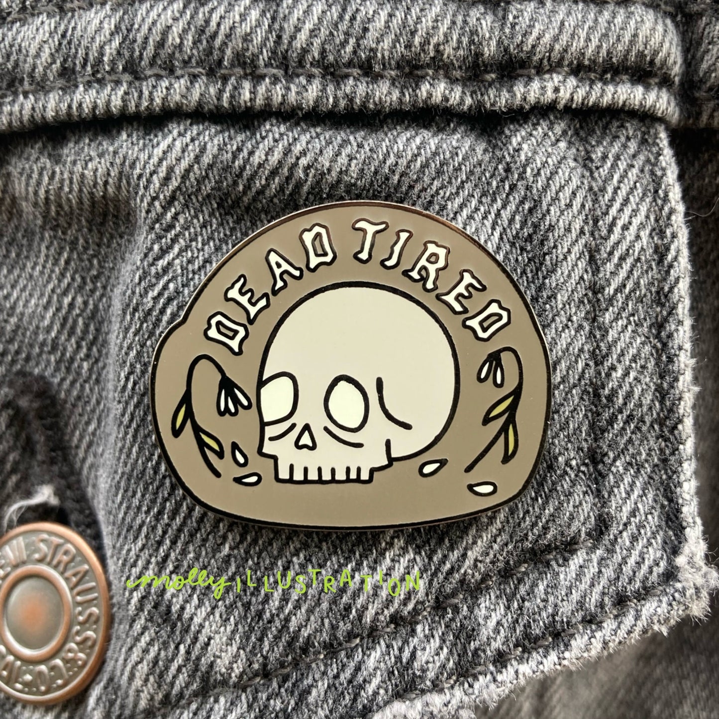 an image of a hard enamel pin that reads dead tired over cartoon skull and dying flowers pinned to a denim jacket.