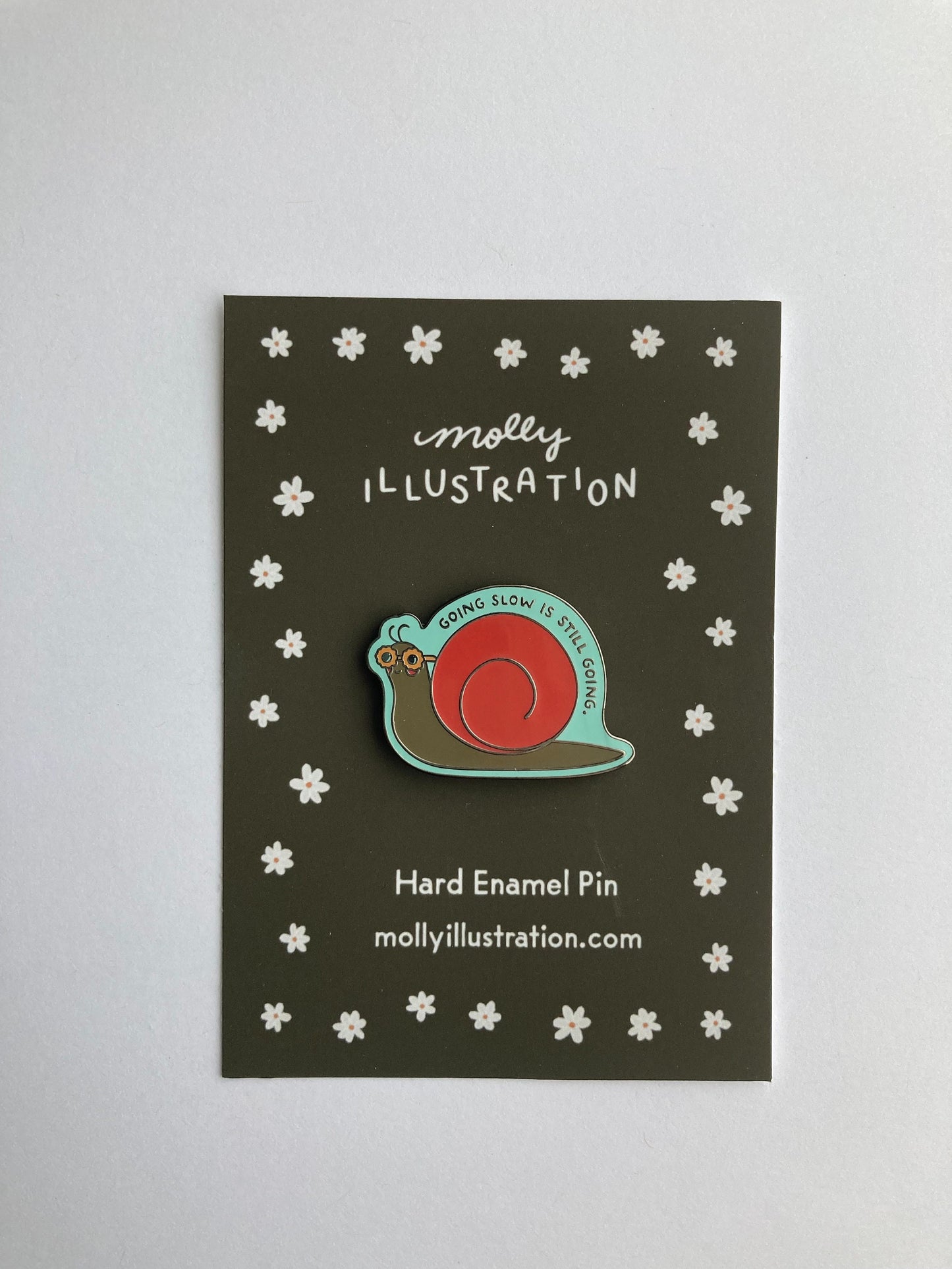 an illustrated hard enamel pin of a snail that is wearing sunglasses. above it reads "going slow is still going"