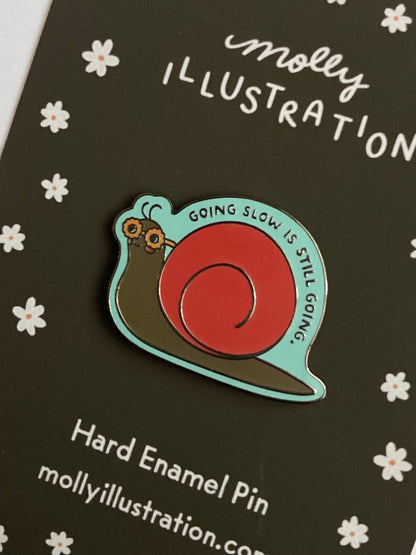 an illustrated hard enamel pin of a snail that is wearing sunglasses. above it reads "going slow is still going"