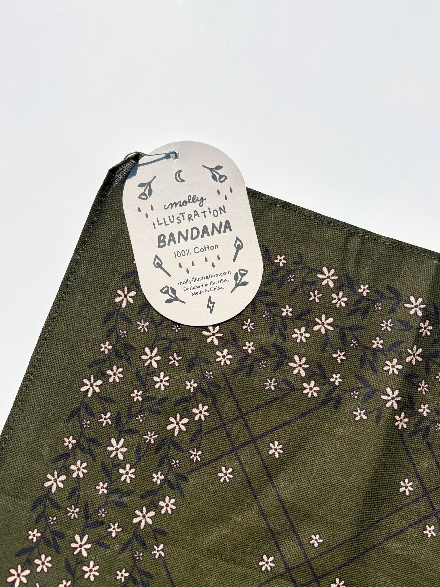 An olive green cotton bandana with a floral pattern, featuring a white paper hangtag that reads 'Molly ILLUSTRATION BANDANA 100% Cotton'.