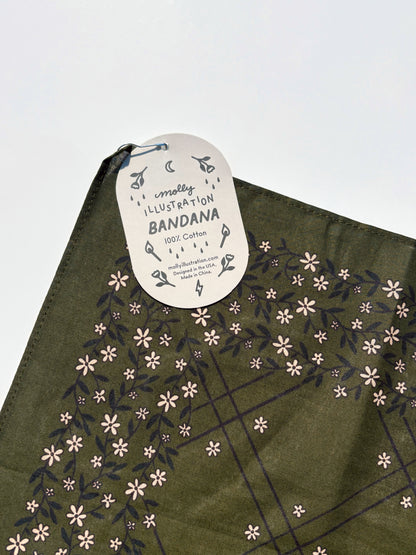 An olive green cotton bandana with a floral pattern, featuring a white paper hangtag that reads 'Molly ILLUSTRATION BANDANA 100% Cotton'.