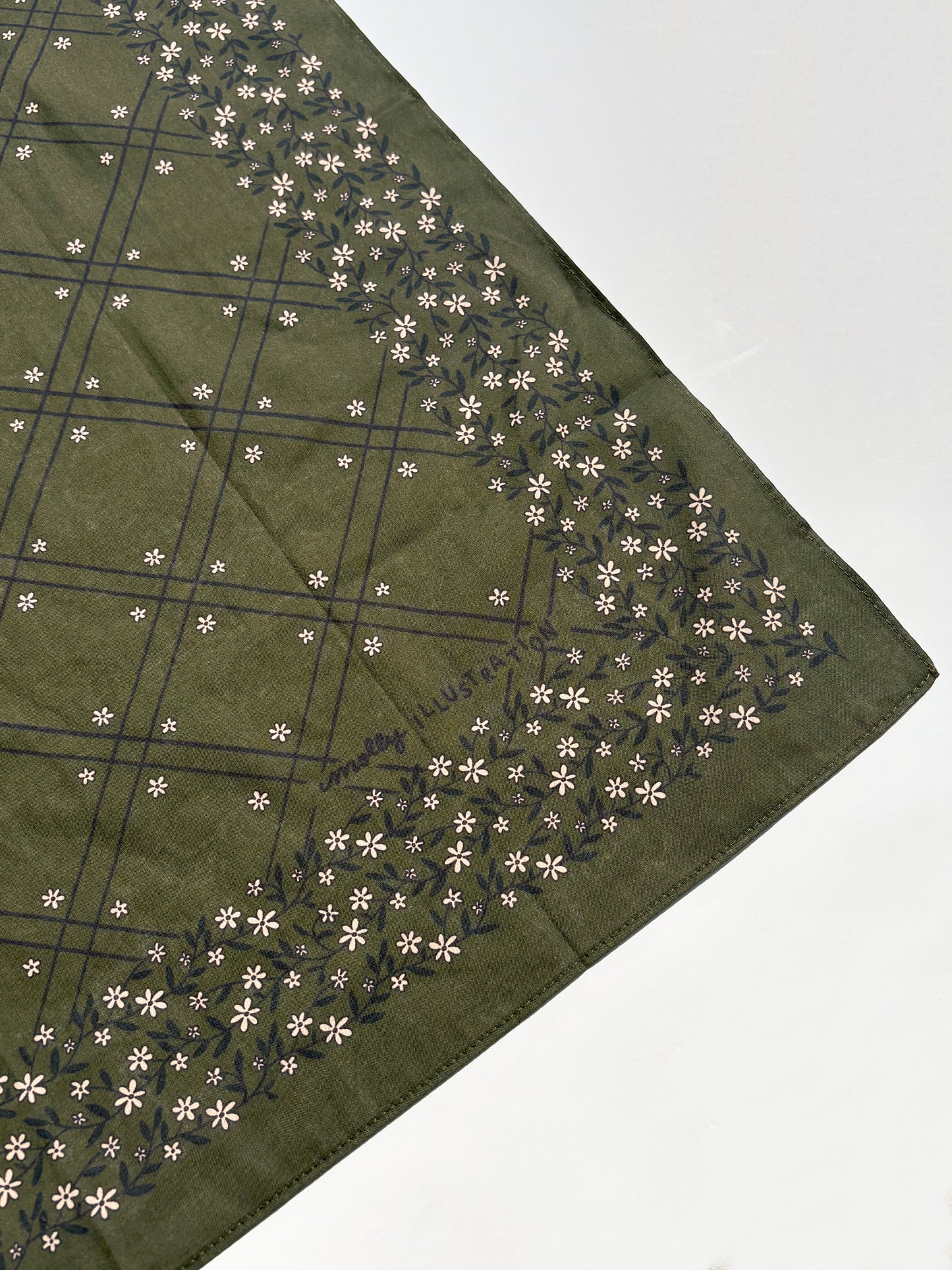 An olive green square scarf with a dark green grid pattern and a border of small white flowers, laid flat on a white surface.