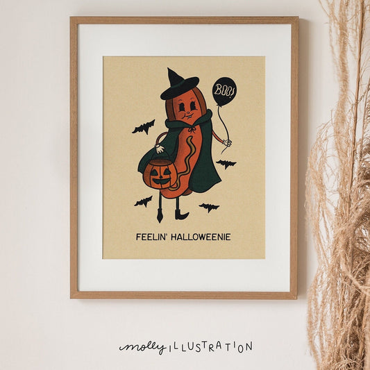 An illustrated art print of a Halloween Hot Dog (halloweenie) dressed as a witch and holding a jack o lantern bucket. Beneath it reads: "Feelin' Halloweenie"