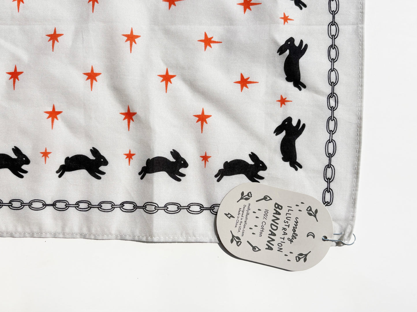 A white fabric bandana with a pattern of black rabbits and orange stars, finished with a black chain border, shown with its paper brand tag.