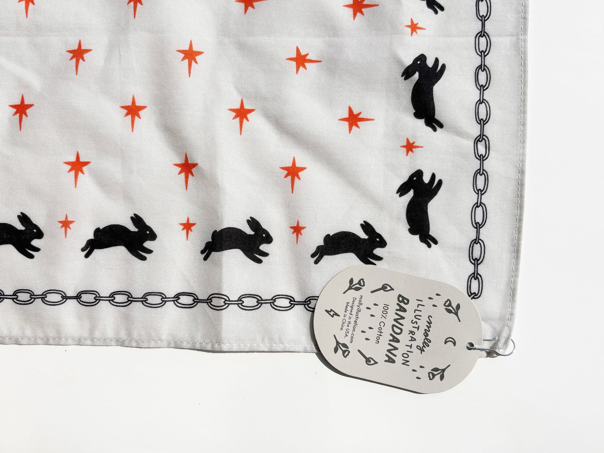 A white fabric bandana with a pattern of black rabbits and orange stars, finished with a black chain border, shown with its paper brand tag.
