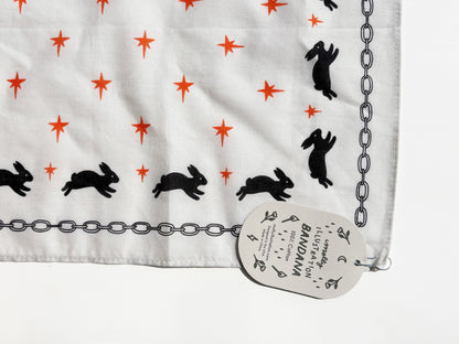 A white fabric bandana with a pattern of black rabbits and orange stars, finished with a black chain border, shown with its paper brand tag.