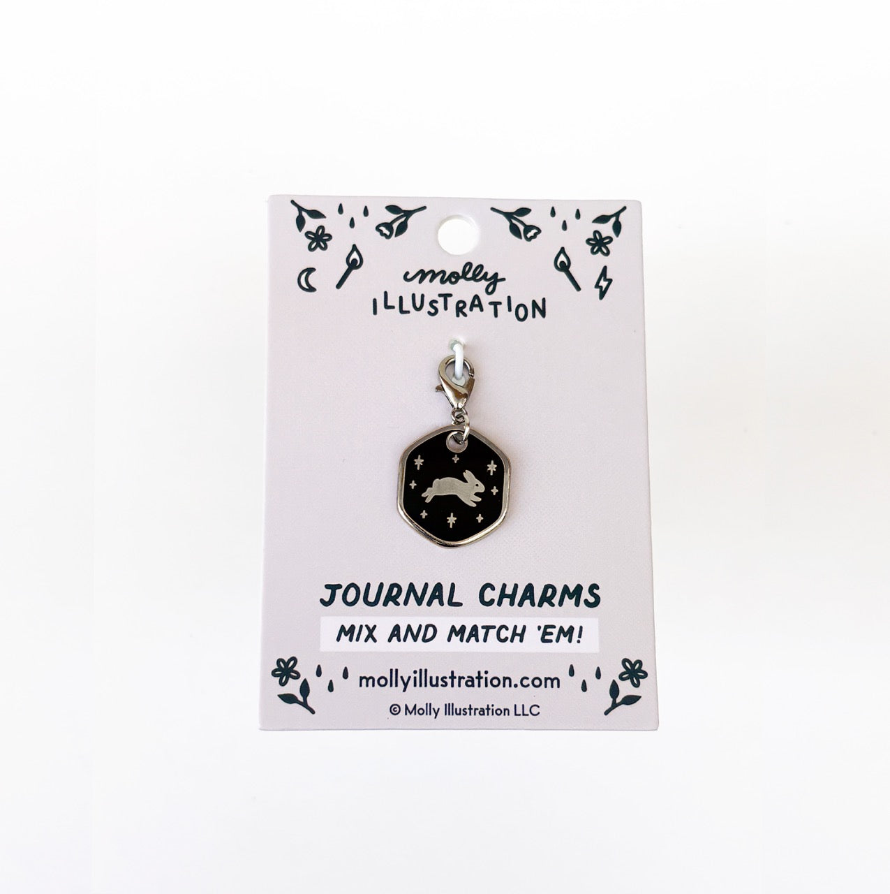 A silver and black journal charm with a leaping bunny on it. Behind it is a branded backer card for the product