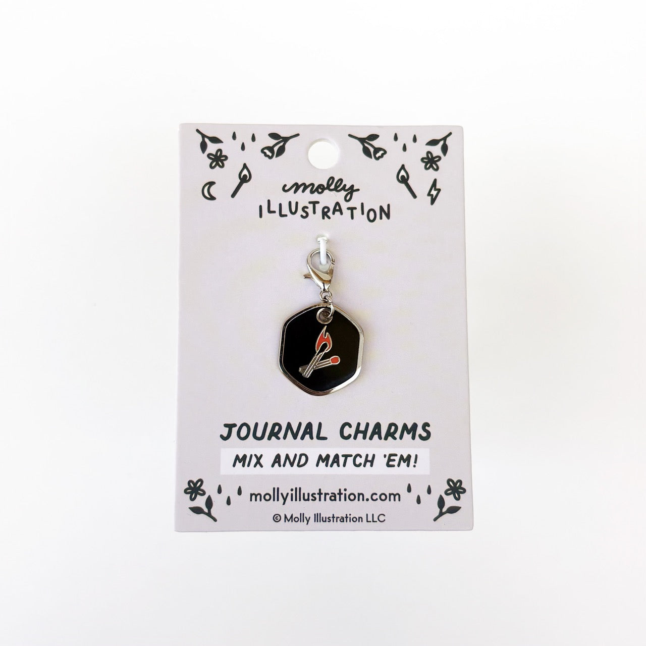 A silver and black journal charm with a two matches on it. Behind it is a branded backer card for the product