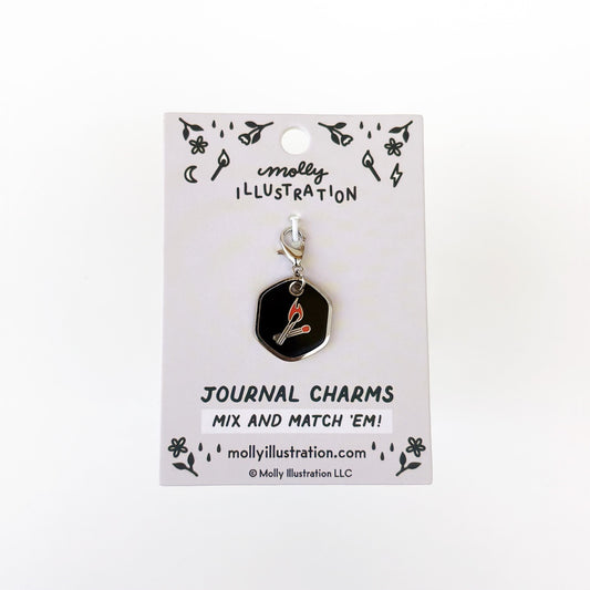 A silver and black journal charm with a two matches on it. Behind it is a branded backer card for the product