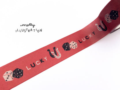 A roll of illustrated washi tape printed with the phrase “Lucky” printed on it alongside dice, and horseshoes.