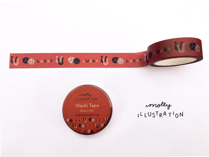 A roll of illustrated washi tape printed with the phrase “Lucky” printed on it alongside dice, and horseshoes.