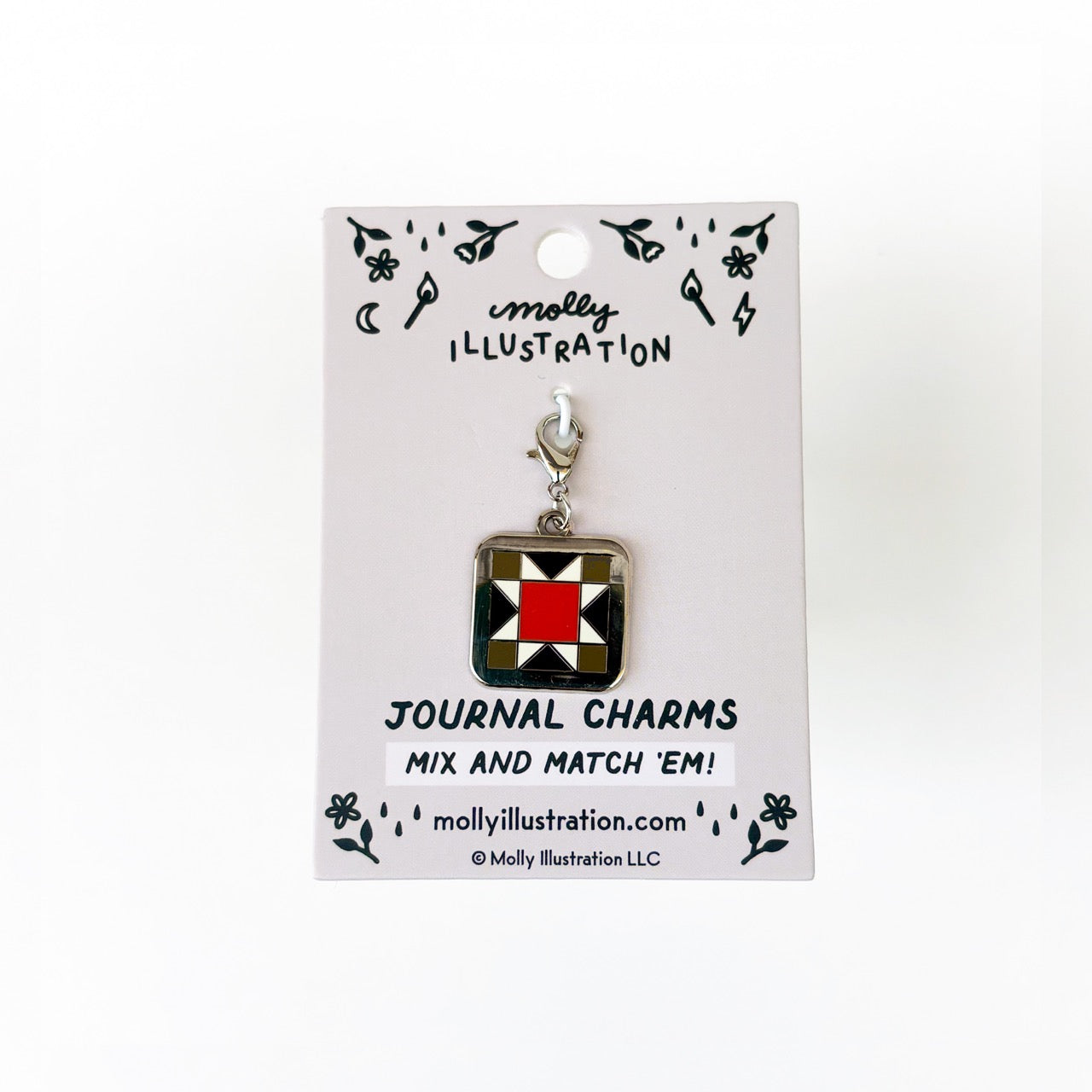 A silver, black,red and green journal charm of a quilt square. Behind it is a branded backer card for the product