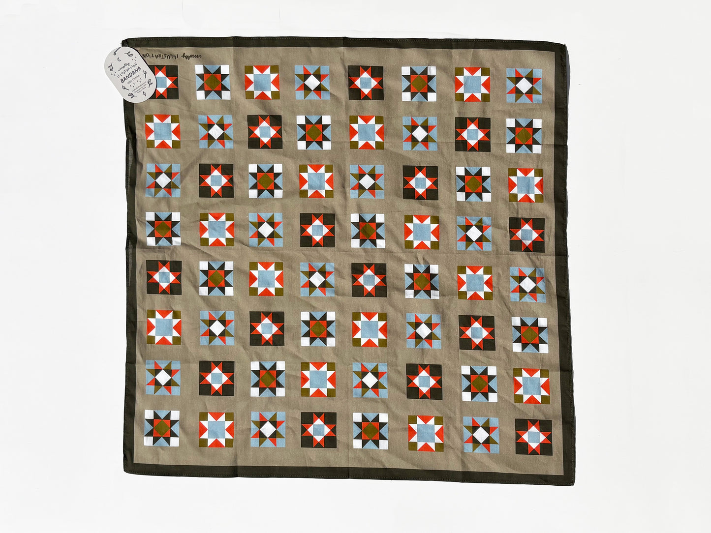 A square quilt laid flat on a white background, featuring a tan center with a grid of geometric star and diamond blocks in red, blue, black, white, and brown, finished with a dark brown border. A small white paper tag is attached at the top left corner.