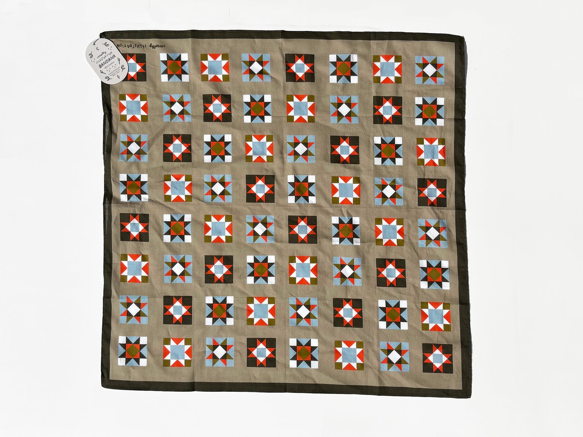 A square quilt laid flat on a white background, featuring a tan center with a grid of geometric star and diamond blocks in red, blue, black, white, and brown, finished with a dark brown border. A small white paper tag is attached at the top left corner.