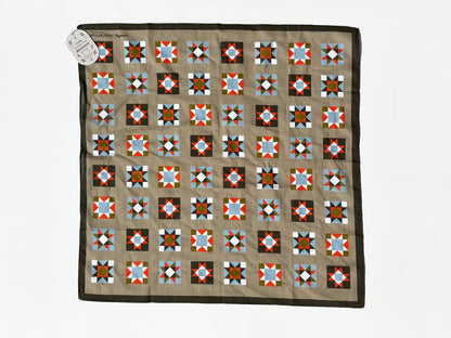 A square quilt laid flat on a white background, featuring a tan center with a grid of geometric star and diamond blocks in red, blue, black, white, and brown, finished with a dark brown border. A small white paper tag is attached at the top left corner.