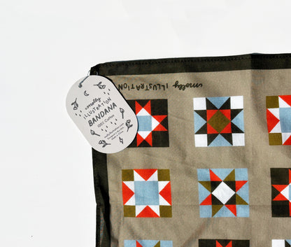 A close-up of a fabric bandana with a geometric quilt pattern, featuring a light brown background and colorful blocks of red, blue, green, and black. A white paper tag is attached to the corner.