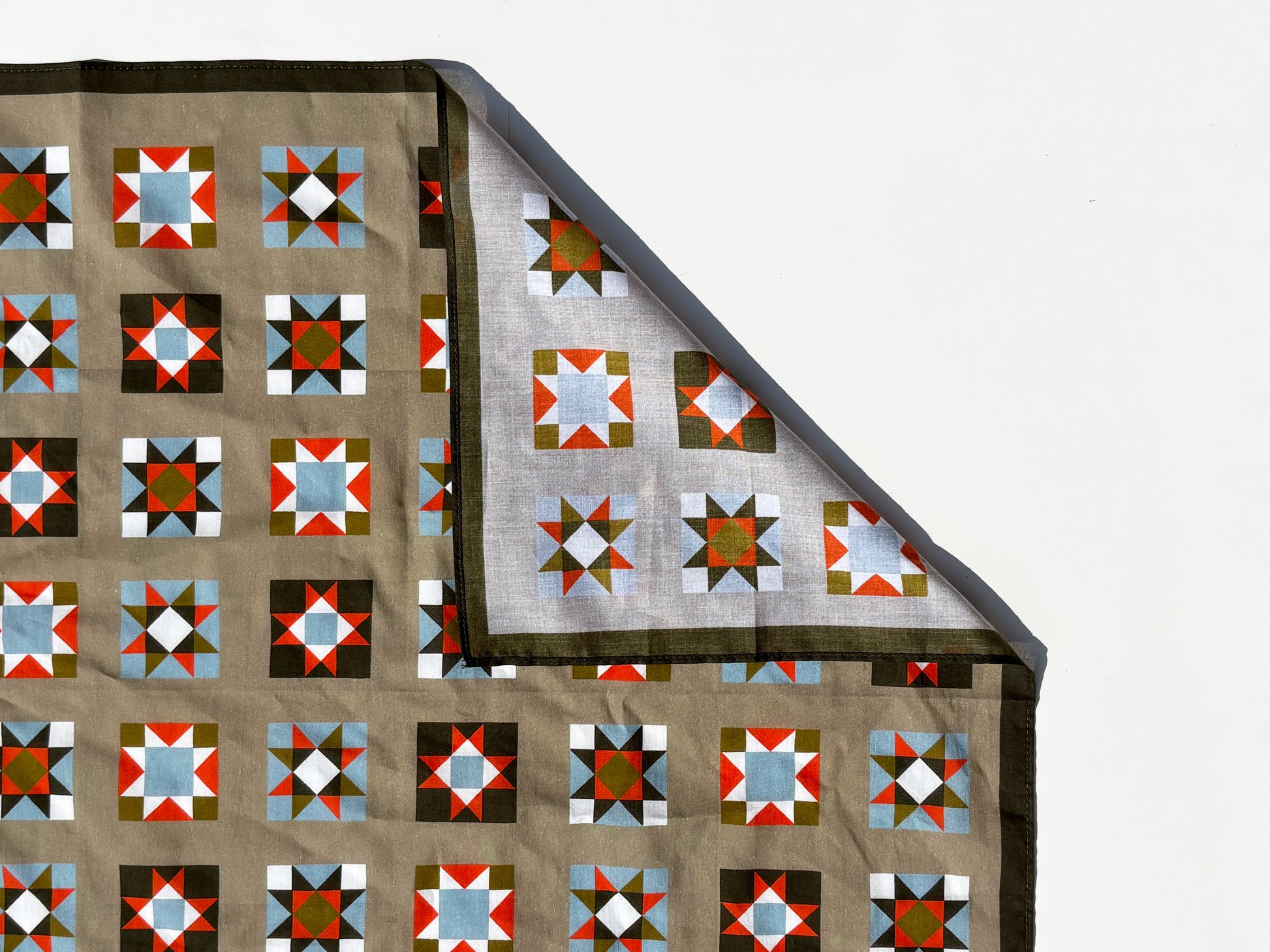 A folded quilt or blanket with a geometric block pattern in brown, red, blue, white, and green, shown against a plain white background with one corner folded over to reveal the reverse side.