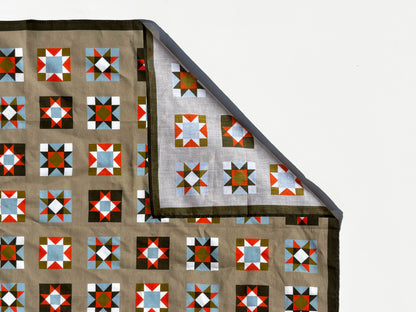 A folded quilt or blanket with a geometric block pattern in brown, red, blue, white, and green, shown against a plain white background with one corner folded over to reveal the reverse side.
